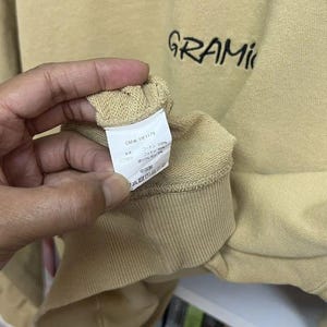 May include: Tan-colored sweatshirt with the word "GRAMIC" embroidered in black. The sleeve cuff is visible, and a tag with text is held by a hand. The sweatshirt appears to be made of a soft material.