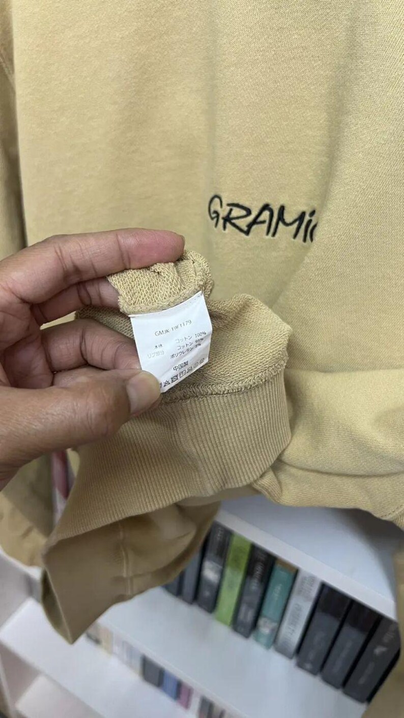 May include: Tan-colored sweatshirt with the word "GRAMIC" embroidered in black. The sleeve cuff is visible, and a tag with text is held by a hand. The sweatshirt appears to be made of a soft material.