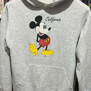 May include: A light grey long-sleeve hooded sweatshirt featuring a graphic of Mickey Mouse in red shorts and yellow shoes. The word California is printed in black script above the character. The garment includes a front kangaroo pocket and a hood.