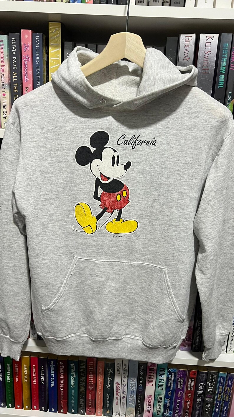 May include: A light grey long-sleeve hooded sweatshirt featuring a graphic of Mickey Mouse in red shorts and yellow shoes. The word California is printed in black script above the character. The garment includes a front kangaroo pocket and a hood.
