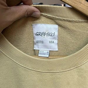 May include: A tan Gramicci sweatshirt with a crew neck. The label reads "GRAMICCI ORIGINAL FREEDOM USA". The size is listed as M for Asia and S for USA/EU. The sweatshirt is hanging on a wooden hanger.