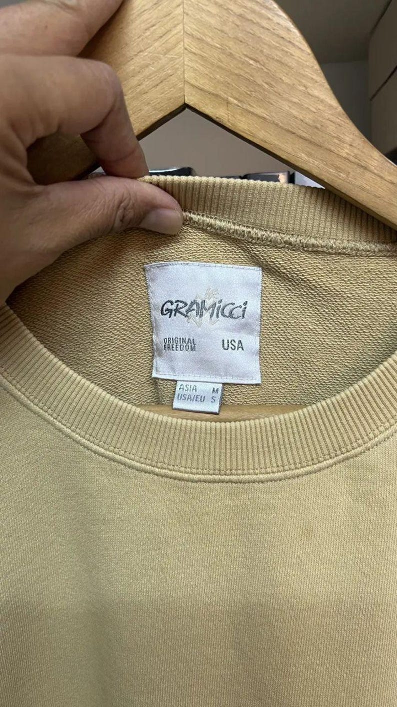 May include: A tan Gramicci sweatshirt with a crew neck. The label reads "GRAMICCI ORIGINAL FREEDOM USA". The size is listed as M for Asia and S for USA/EU. The sweatshirt is hanging on a wooden hanger.