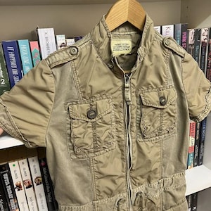 Puede incluir: Una chaqueta utilitaria de color caqui de manga corta con cremallera frontal completa, cuello alzado y cuatro bolsillos con solapa y bot&oacute;n. La chaqueta presenta charreteras en los hombros, cord&oacute;n en la cintura y etiqueta AVIREX LTD en el cuello.