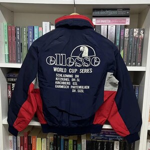 Puede incluir: Una chaqueta azul marino con detalles en rojo y gris en las mangas y cuello alto. La parte trasera tiene texto blanco que dice ellesse WORLD CUP SERIES, enumerando lugares como SCHLADMING, KITZBÜHEL, KIRCHBERG y GARMISCH PARTENKIRCHEN.