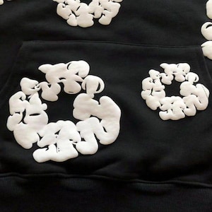 May include: Black hooded sweatshirt with white, cloud-like, puffy designs. The sweatshirt features a front pocket and the cloud designs are placed on the hood and front of the sweatshirt. The design is a unique, modern take on a classic hoodie.