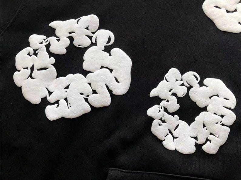 May include: Black hoodie featuring white, cloud-like embroidered designs. The soft, textured white shapes are arranged in circular patterns across the dark fabric, creating a striking contrast. The hoodie has a front pocket.