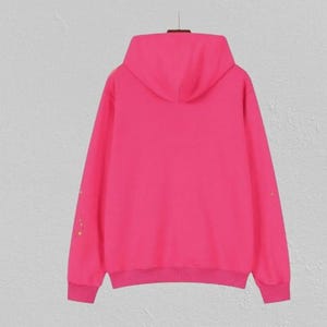 May include: A bright pink hoodie with a matching hood and ribbed cuffs and hem. The long sleeves have a subtle design of small, colourful embellishments. The hoodie is displayed on a hanger against a neutral background.
