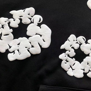 May include: Black hoodie featuring white, cloud-like embroidered designs. The design is circular and abstract, with a textured appearance. The hoodie has a simple, minimalist aesthetic, with a focus on the contrasting colors and shapes.