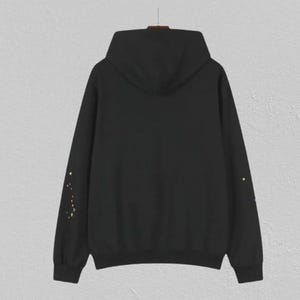May include: Black hoodie with a hood and long sleeves. The sleeves feature a constellation design in yellow and gold. The hoodie is hanging on a hanger against a neutral background. A casual, comfortable garment.