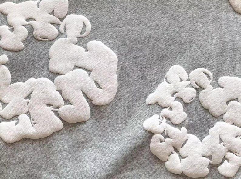 May include: Close-up of a gray fabric with several white, cloud-shaped appliqu&eacute;s. The appliqu&eacute;s have a textured surface and are arranged in clusters across the fabric. The shapes have rounded edges and a soft appearance.