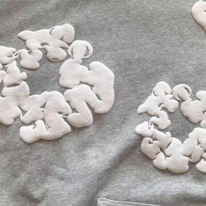 May include: Close-up of a gray hoodie with white, cloud-shaped embroidered designs. The soft, textured fabric features multiple cloud clusters, creating a unique and artistic pattern. The hoodie's design is simple yet eye-catching.