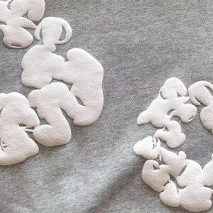 May include: Close-up of a gray fabric with multiple white, cloud-shaped appliqu&eacute;s. The appliqu&eacute;s are textured and arranged in clusters across the surface, creating a decorative pattern. The fabric appears to be a soft material.