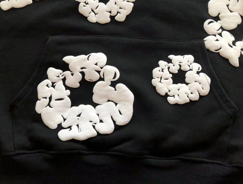 May include: Black hoodie with white, cloud-like, textured designs. The hoodie features a front pocket and a drawstring hood. The white designs are clustered in circular patterns across the front and on the hood.