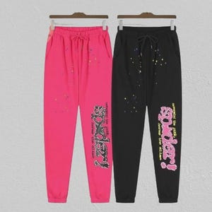 May include: Two pairs of sweatpants, one hot pink and one black, hanging on a rack. Both pairs have a drawstring waist and a graphic that says "Spider!" in a stylized font. The pink pair has colourful paint splatters.