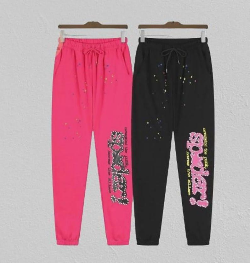 May include: Two pairs of sweatpants, one hot pink and one black, hanging on a rack. Both pairs have a drawstring waist and a graphic that says "Spider!" in a stylized font. The pink pair has colourful paint splatters.