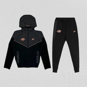 Pode incluir: Conjunto de fato de treino preto com casaco com capuz e fecho de correr e calças a condizer. O casaco apresenta uma faixa e um logótipo dourados, com um logótipo pequeno no peito. As calças têm cintura com cordão e um logótipo dourado. O conjunto é feito de material macio e confortável.