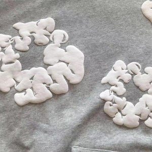 May include: Close-up of a gray hoodie with white, cloud-shaped appliqu&eacute;s. The appliqu&eacute;s are arranged in circular patterns. The fabric has a soft, textured appearance. The design is simple and modern.