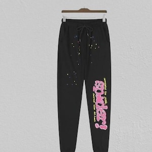 May include: Black sweatpants with a drawstring waist and pockets. The trousers have colourful paint splatter details and the text "powder!" in pink and yellow, with additional text below.