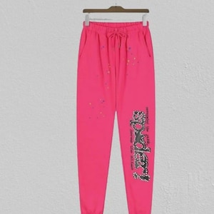 May include: Vibrant pink sweatpants with a drawstring waist and pockets. The trousers have a rhinestone design on the left leg with the word "Bodery" and colourful speckles. They are hanging on a wooden hanger.