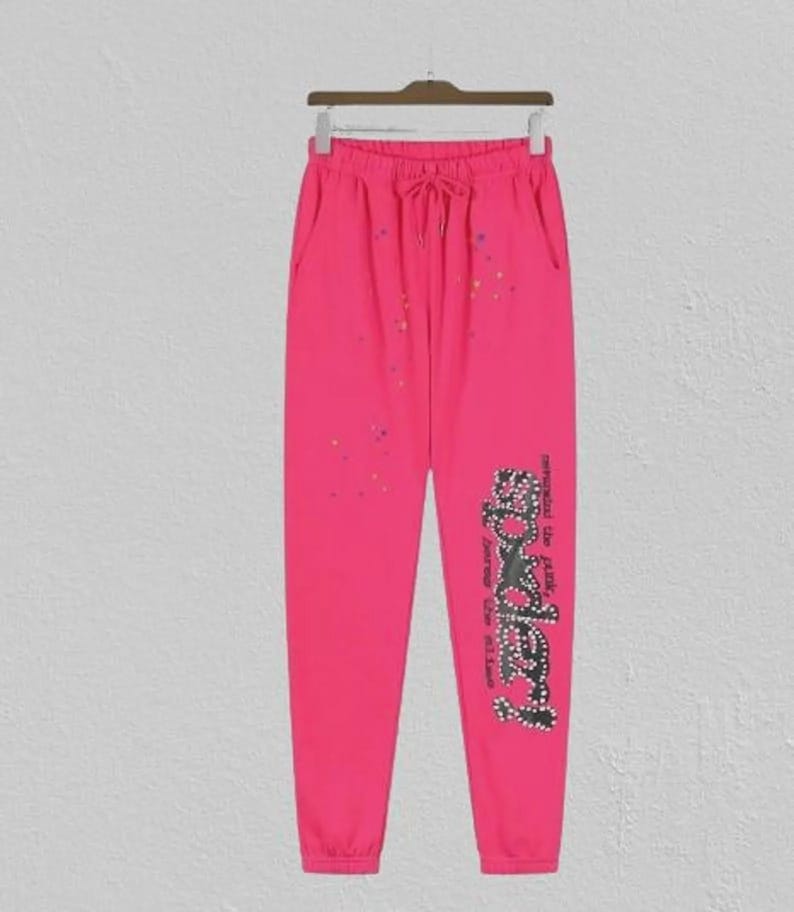 May include: Vibrant pink sweatpants with a drawstring waist and pockets. The trousers have a rhinestone design on the left leg with the word "Bodery" and colourful speckles. They are hanging on a wooden hanger.