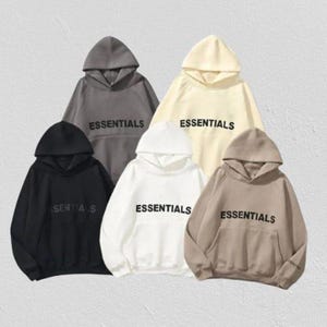 Op de afbeelding: Vijf hoodies in verschillende kleuren: zwart, grijs, wit, beige en bruin. Elke hoodie heeft de tekst "ESSENTIALS" op de borst. De hoodies worden gepresenteerd tegen een neutrale achtergrond.