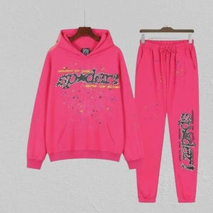 May include: A vibrant pink tracksuit comprising a hooded sweatshirt and joggers. The sweatshirt displays the word "SPYDER" in a decorative font across the chest. The joggers have a drawstring waist and the same "SPYDER" design on the leg.
