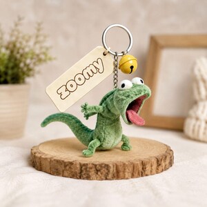 May include: A green plush lizard keyring with an open mouth and pink tongue. The keyring has a wooden tag with "Zoomy" and a small yellow bell. The lizard is displayed on a wooden slice.