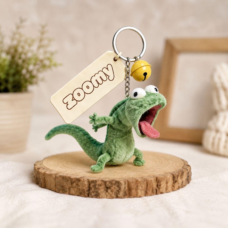 May include: A green plush lizard keyring with an open mouth and pink tongue. The keyring has a wooden tag with "Zoomy" and a small yellow bell. The lizard is displayed on a wooden slice.