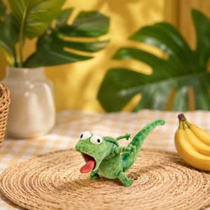 May include: A green plush chameleon toy with a red tongue, on a woven mat. The toy has large eyes and outstretched arms. A bunch of bananas is in the background. The scene is set on a yellow and white checkered tablecloth.