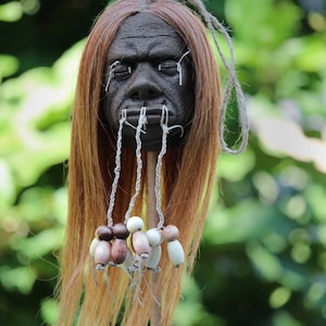May include: A dark-skinned human head with long brown hair, sewn shut eyes, and a mouth sewn shut with white thread. The head is on a wooden stand with white and brown beads hanging from the mouth.