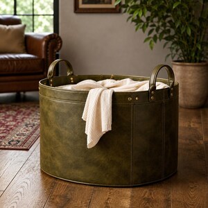 May include: A round, olive green storage basket with two matching handles attached by brass-coloured rivets. The basket features a smooth, textured exterior and contains a folded, cream-coloured knit textile draped over the top rim.