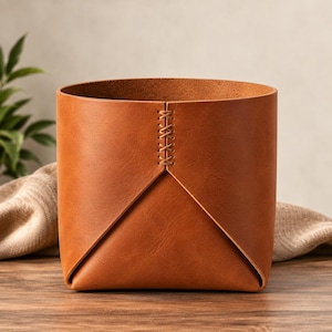Pode incluir: Um recipiente de armazenamento cilíndrico feito de couro liso castanho-claro. O exterior apresenta uma costura vertical proeminente unida por um padrão decorativo de ponto cruz, com o couro dobrado numa forma triangular na base.
