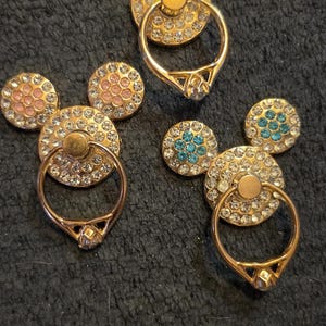 Peut inclure: Supports de bague de t&eacute;l&eacute;phone dor&eacute;s en forme de Mickey Mouse. Chaque support est dot&eacute; d'une bague pour la prise en main et est orn&eacute; de strass. Les accents d'oreille sont rouges, roses et turquoise. Ces accessoires ajoutent une touche d'&eacute;clat et de fun &agrave; tout t&eacute;l&eacute;phone.
