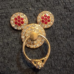 Peut inclure: Un support de bague de t&eacute;l&eacute;phone dor&eacute; en forme de Mickey Mouse. Les oreilles sont rondes, orn&eacute;es de strass clairs et rouges. Le cercle central a une bague attach&eacute;e, &eacute;galement dor&eacute;e, avec un strass clair.