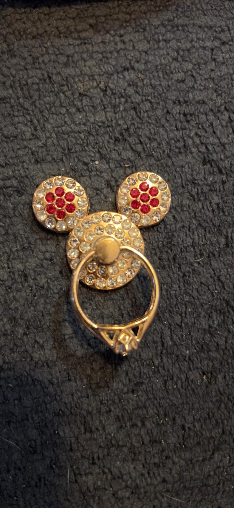 Peut inclure: Un support de bague de t&eacute;l&eacute;phone dor&eacute; en forme de Mickey Mouse. Les oreilles sont rondes, orn&eacute;es de strass clairs et rouges. Le cercle central a une bague attach&eacute;e, &eacute;galement dor&eacute;e, avec un strass clair.