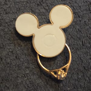Peut inclure: Un support de t&eacute;l&eacute;phone en forme de Mickey Mouse blanc et dor&eacute;, avec un anneau dor&eacute; et une gemme transparente. La poign&eacute;e est con&ccedil;ue pour se fixer au dos d'un t&eacute;l&eacute;phone.