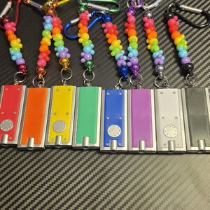 May include: Eight rectangular flashlights with metallic silver bodies and colored panels in red, orange, yellow, green, blue, purple, white, and black. Each flashlight features a circular button and a rainbow-colored beaded lanyard with a metal carabiner.