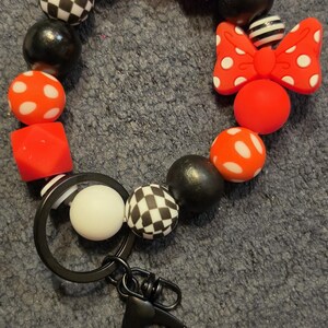 May include: A circular keychain made of silicone beads in red, white, and black. The beads feature solid colors, white polka dots on red, black and white checkerboard patterns, and a red bow with white polka dots. A black metal ring and clasp are attached.