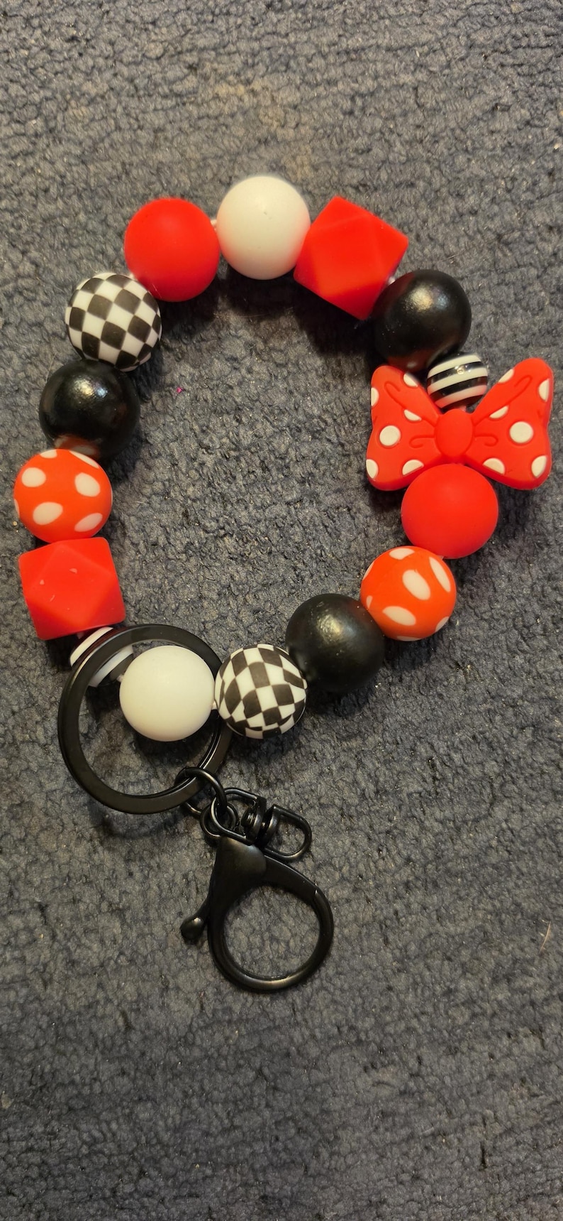 May include: A circular keychain made of silicone beads in red, white, and black. The beads feature solid colors, white polka dots on red, black and white checkerboard patterns, and a red bow with white polka dots. A black metal ring and clasp are attached.