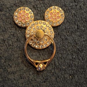 Peut inclure: Un support de bague de t&eacute;l&eacute;phone dor&eacute; en forme de t&ecirc;te de Mickey Mouse. Les trois formes circulaires sont orn&eacute;es de strass transparents et roses. La bague est dor&eacute;e et poss&egrave;de un accent en forme de diamant.