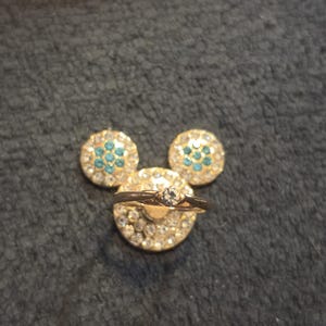 Peut inclure: Une bague dor&eacute;e avec un diamant central repose sur une base dor&eacute;e recouverte de strass, flanqu&eacute;e de deux boucles d'oreilles assorties en strass et turquoise, dispos&eacute;es en forme de Mickey Mouse.