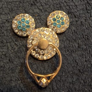 Peut inclure: Un support de bague de t&eacute;l&eacute;phone dor&eacute; en forme de Mickey Mouse. Le cercle central et les deux cercles en forme d'oreille sont incrust&eacute;s de strass transparents et bleus. La bague est dor&eacute;e et poss&egrave;de un accent en forme de diamant.