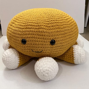 May include: A large, round, crocheted octopus in shades of mustard yellow and white. The octopus has black button eyes and a smiling mouth. The tentacles are white with yellow tips. This handmade plush toy is a whimsical addition to any room.
