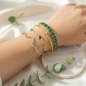 Peut inclure: Plusieurs bracelets dorés ornent un poignet. On distingue un jonc en or orné d'émeraudes, une fine chaîne avec des accents verts émeraude et un bracelet ondulé avec des cristaux transparents. Un bracelet en or avec une pierre transparente est également visible.