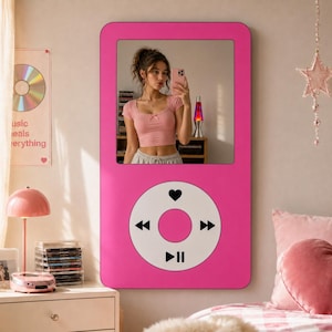 May include: A large, pink mirror in the shape of a classic music player. The mirror has a rectangular frame with rounded corners and a white circular control panel with black icons. The mirror reflects a person taking a selfie.