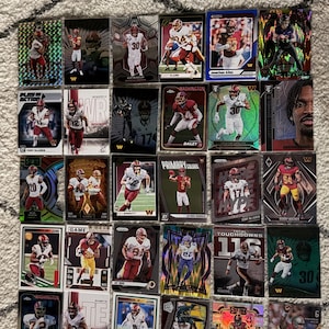 May include: A collection of twenty-eight Washington Commanders football trading cards arranged in a grid. Each card features a player in uniform, with various holographic, metallic, and graphic designs, including text like Play Action, Primary Colors, and Sunday Swag.
