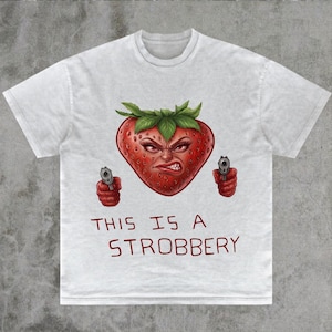 May include: White t-shirt featuring a red strawberry with an angry face, green leaves, and two hands holding guns. The text "THIS IS A STROBBERY" is printed below the graphic.