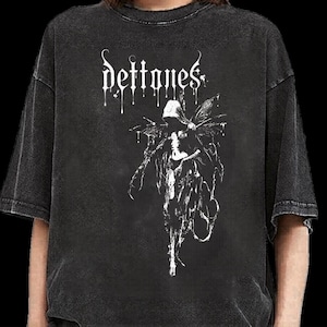 May include: A charcoal grey short-sleeve t-shirt featuring a white graphic print on the chest. The design displays the text "deftones" in a stylized dripping font above a detailed illustration of a hooded figure with wings and flowing robes.