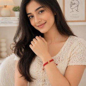 Könnte beinhalten: Ein rotes geflochtenes Armband mit einem goldenen Buchstaben "A" Anhänger. Das Armband wird am Handgelenk einer Person getragen. Die Person trägt ein cremefarbenes Oberteil mit offener Strickstruktur. Der Hintergrund umfasst eine Lampe, Bücher und einen gerahmten Matisse-Druck.