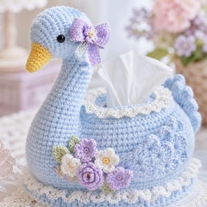 May include: A light blue crocheted swan tissue box cover with a yellow beak and a purple bow. The swan is decorated with white and purple crocheted flowers and a white lace trim. Tissue papers are visible.